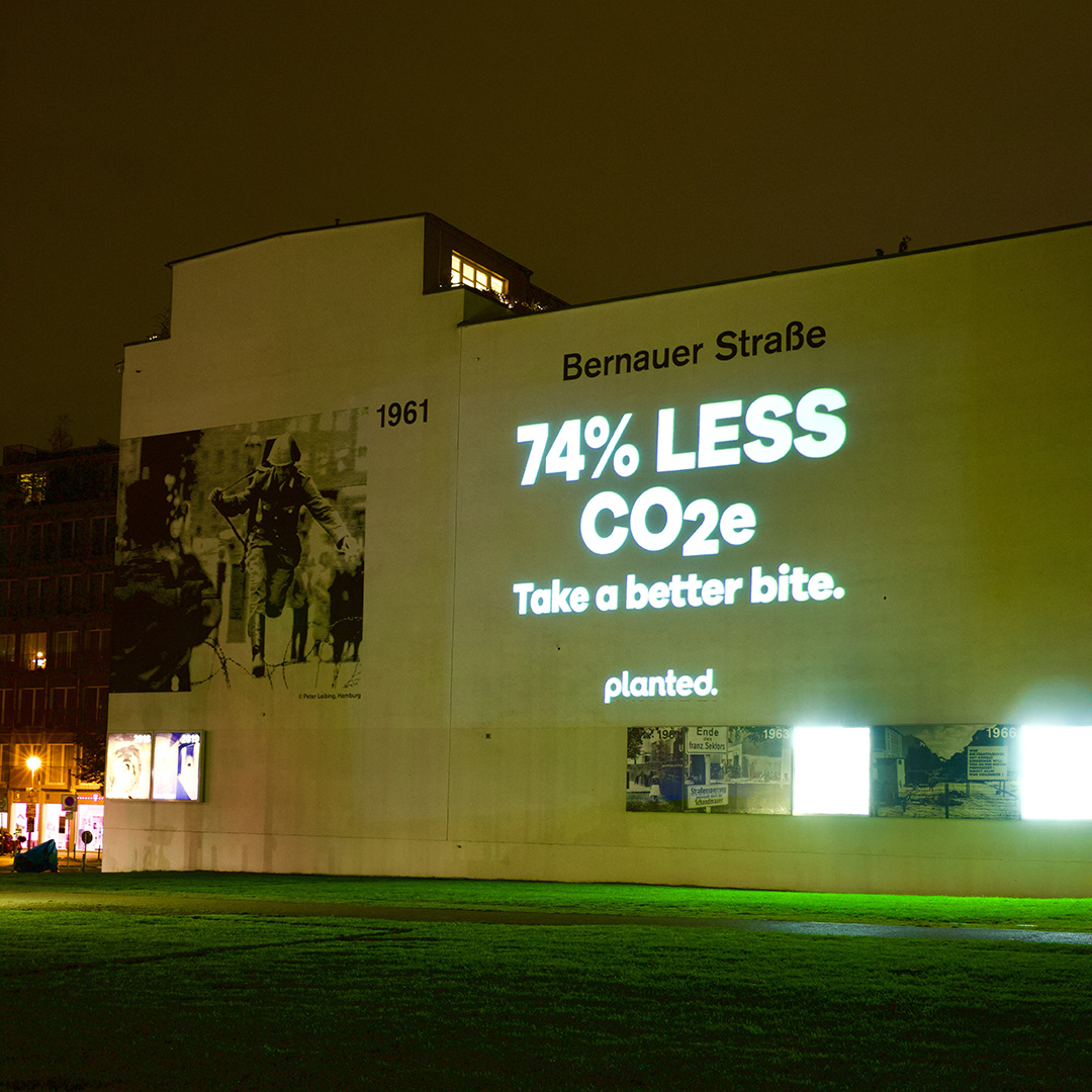 ‘74% less CO₂e. Take a better bite.’ projected onto a wall on Bernauer Straße
